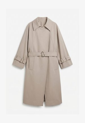 Trench-coat long beige avec col large, taille cintrée par une ceinture à boucle métallique unique, et poignets assortis ceinturés avec garniture métallique.