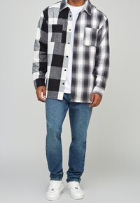 Camisa de franela de cuadros en blanco y negro, con un diseño de botones, que presenta un solo bolsillo en el pecho. Usada sobre una camiseta blanca y jeans azules.