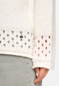 Cremfarbener Strickpullover mit strukturiertem Muster, der über ein Lochmuster an den Bündchen verfügt. Silberne Metallakzente in der Nähe des Saums.