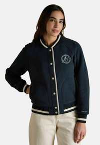 Tumesin fleece-jakk valgete aktsentidega, soonikute äärtega ja rinnal logoga. Omab nuppe ja küljetaskuid. Sile tekstuur.