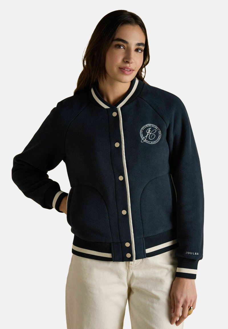 Tumesin fleece-jakk valgete aktsentidega, soonikute äärtega ja rinnal logoga. Omab nuppe ja küljetaskuid. Sile tekstuur.