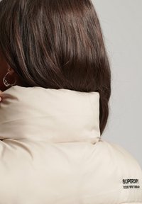 Beige vadderad jackkrage med texturerad yta, med svart broderi av "SUPERDRY CODE SPORTSWEAR", sedd bakifrån.