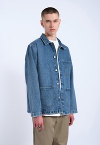 Giacca di denim leggera di colore blu chiaro con chiusura a bottoni, due tasche frontali e colletto abbattuto. Presenta una vestibilità rilassata e maniche risvoltate.