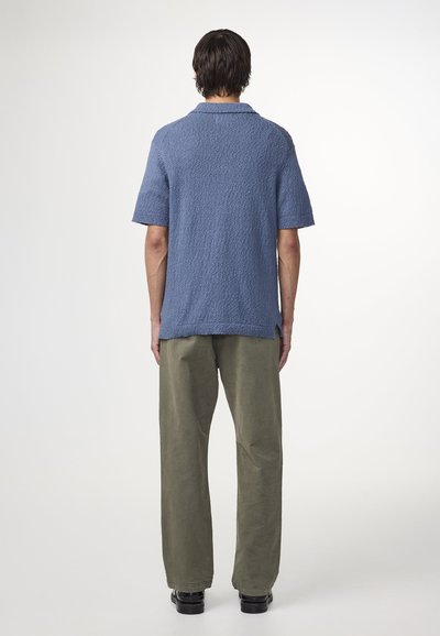 Camicetta corta blu a trama con colletto, abbinata a pantaloni verdi larghi. L'outfit ha una vestibilità rilassata e dettagli minimi.