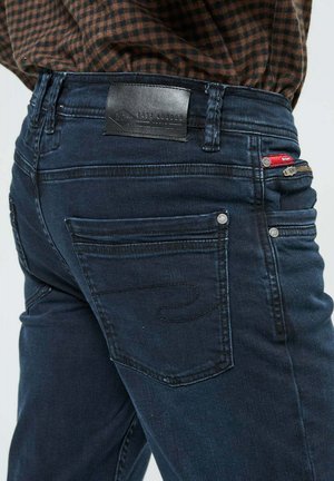 Jean en denim foncé avec un design ajusté, comportant une étiquette en cuir à la taille, plusieurs poches et des détails de couture discrets.