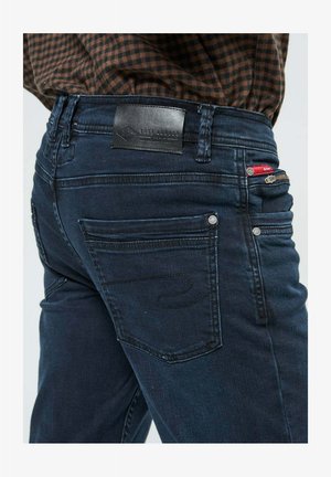 Jean en denim foncé avec un design ajusté, comportant une étiquette en cuir à la taille, plusieurs poches et des détails de couture discrets.