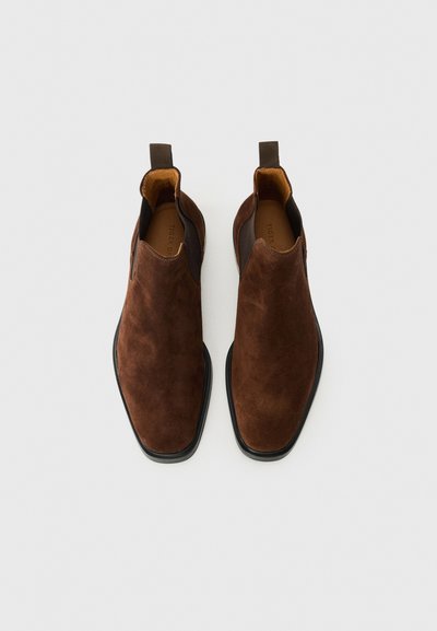 O pereche de cizme Chelsea din suede maro, cu panouri laterale elastice negre și tălpi de cauciuc negre, văzute din perspectiva de sus pe un fundal alb.