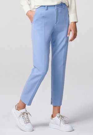 Femme portant un pantalon court ajusté bleu clair avec des plis avant et des baskets blanches avec des accents dorés, debout avec une main dans la poche.