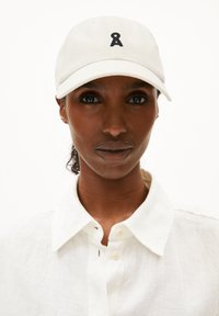 Witte baseballpet met een zwart geborduurd logo, gemaakt van katoen, met een gebogen klep en een verstelbare strap aan de achterkant, gedragen met een wit overhemd met kraag.