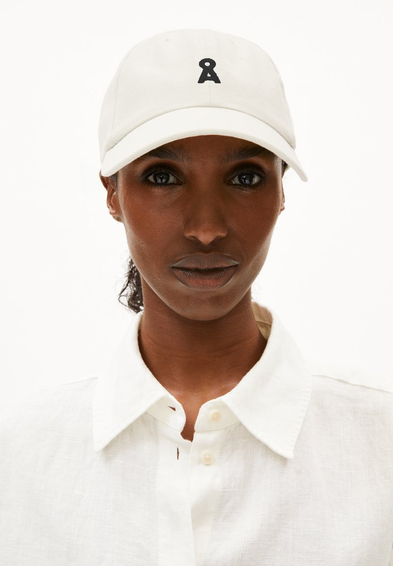 Witte baseballpet met een zwart geborduurd logo, gemaakt van katoen, met een gebogen klep en een verstelbare strap aan de achterkant, gedragen met een wit overhemd met kraag.