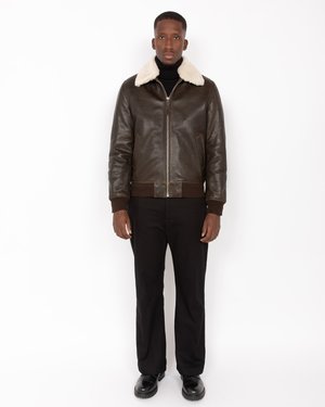 Chaqueta de cuero marrón con un cuello de borrego crema, cierre de cremallera en la parte frontal y puños acanalados. Combinada con pantalones y zapatos negros.
