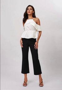 Haut peplum blanc décolleté avec des accents volants associé à un pantalon noir court. Le mannequin porte des talons noirs à lanières. Texture de tissu lisse.
