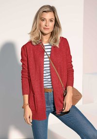 Rote Strickjacke mit gerippten Bündchen und Taschen, kombiniert mit einem gestreiften Hemd. Das Model trägt Jeans und hält eine Sonnenbrille, dazu eine beige Umhängetasche.