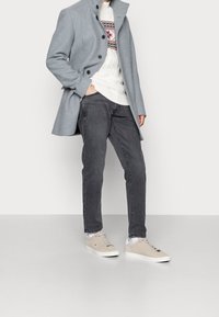 Muž v světle šedém kabátu, bílém vzorovaném svetru, tmavě šedých džínách a béžových teniskách stojí s jednou rukou v kapse.