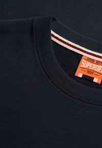 Mørkeblå crewneck sweatshirt med ribbet krave, med et orange "Superdry Goods Dept" mærke og hvide og røde stribede detaljer på den indvendige krave.