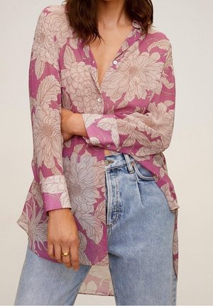 Blouse légère et transparente de couleur rose avec un motif floral et des manches longues. Elle présente une fermeture à boutons et des fentes sur les côtés, portée avec un jean taille haute.