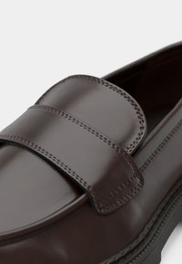 Καφέ δερμάτινο μοκασίνι με λείο φινίρισμα, στρογγυλεμένο σχεδιασμό, ραμμένες λεπτομέρειες και φαρδιά λουρίδα στην κορυφή.