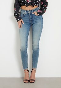 Žena v bledě modrých úzkých džínách, černých sandálech na vysokém podpatku a černém květinovém crop topu s puntíky a dlouhým rukávem.