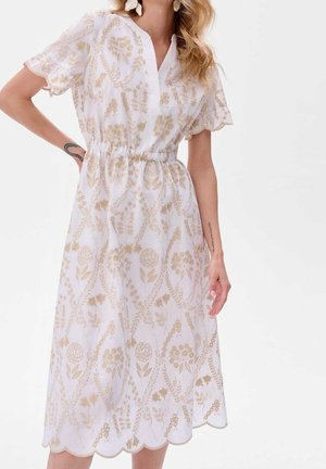 Kvinde iført en hvid midi-kjole med beige blomsterbroderi, korte bølgede ærmer, elastisk talje og V-formet halsudskæring, poserer med den ene hånd på hoften.