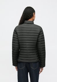 Femeie cu părul lung și ondulat, îmbrăcată într-o jachetă neagră matlasată și blugi de culoare albastru închis, stând cu spatele contra unui fundal simplu.