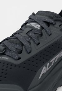 Chaussure de course noire avec une tige texturée, lacets gris et marquage blanc. Présente des perforations pour la respirabilité et une semelle rembourrée.