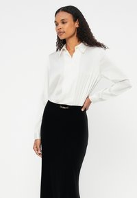 Blusa branca de mangas compridas com colarinho abotoado, combinada com uma saia preta ajustada que apresenta um detalhe de fivela em tom dourado. Tecido suave, design minimalista.