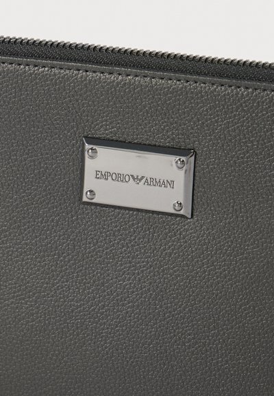 Κοντινή λήψη υφής δερμάτινου θήκης σε σκούρο γκρι χρώμα με ασημένια μεταλλική πλάκα χαραγμένη με το λογότυπο Emporio Armani και μαύρο φερμουάρ στην πάνω άκρη.