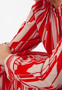 Personne portant une chemise à manches longues rouge et beige avec des rayures abstraites, les bras croisés, sur un fond blanc uni.