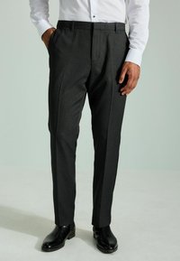 Dunkelgraue maßgeschneiderte Hose mit strukturierter Oberfläche, flachem Vorderteil, seitlichen Taschen und geradem Bein, kombiniert mit schwarzen Schuhen.