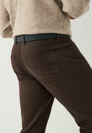 Braune Hosen aus einem robusten Stoff, mit zwei Gesäßtaschen, kontrastierenden Nähten und einem schmalen Schnitt, kombiniert mit einem schwarzen Gürtel.