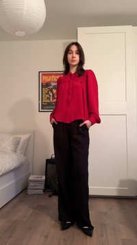 Röd långärmad blus med rynkad halsringning kombinerad med svarta vida byxor. Mjukt tyg, framfickor synliga, stående pose.