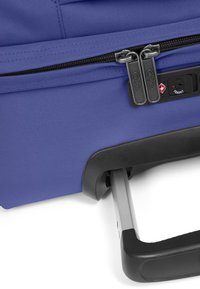 Lila rullväska med texturerad tyg, dubbla blixtlås och ett utdragbart aluminiumhandtag med svart grepp.