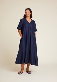 Mørkeblå lin kjole med V-hals, korte rullede ermer, løs passform og sidelommer, som når ned til midten av leggen. Kombinert med blå flip-flops.