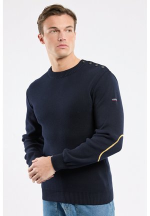 Homme portant un pull bleu marine avec des boutons aux épaules et un patch jaune au coude, associé à un jean bleu clair, regardant sur le côté.