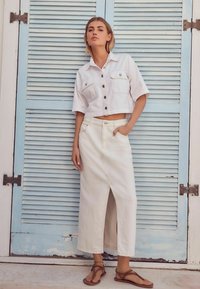 Vrouw in een witte cropped button-up shirt, lange witte rok met een voorklepschaar, en bruine sandalen, staande tegen blauwe luiken.