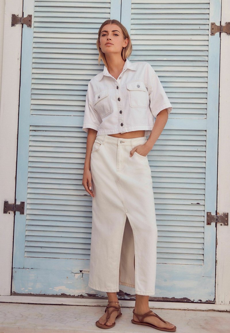Vrouw in een witte cropped button-up shirt, lange witte rok met een voorklepschaar, en bruine sandalen, staande tegen blauwe luiken.