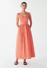 Žena u koraljnoj bezrukavnoj maksi haljini s kvadratnim izrezom, ruku u džepovima, nosi zlatne okrugle naušnice i crne sandale na bijeloj pozadini.