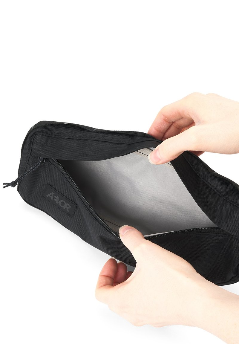 Schwarze Stofftasche mit Reißverschluss, die ein graues Innenfutter zeigt. Eine Hand hält die Tasche und zeigt ein offenes Fach sowie das Markenlogo an der Seite.