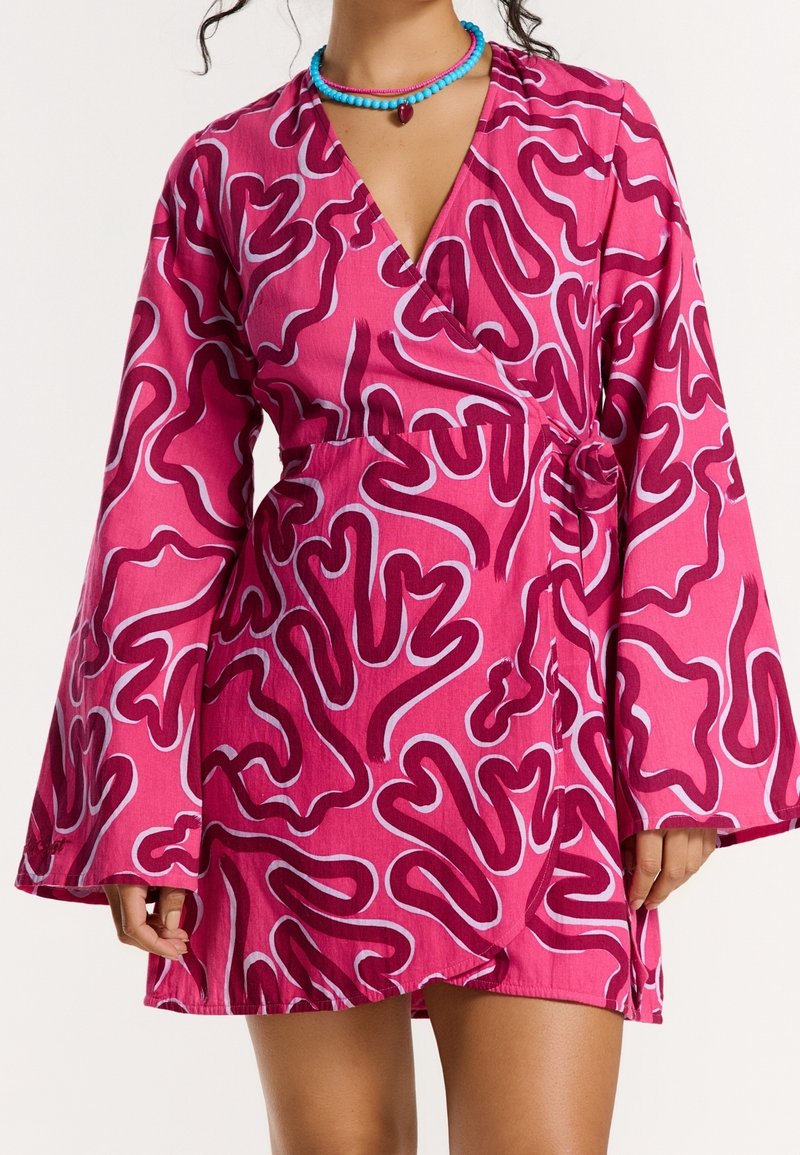 Femme portant une robe portefeuille rose avec des motifs abstraits en zigzag rose foncé et blanc, et des manches évasées larges, accessoirisée de colliers de perles bleues et roses.