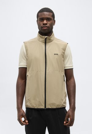 Homme portant un gilet sans manches beige zippé par-dessus une chemise crème à manches courtes, debout devant un fond clair uni.
