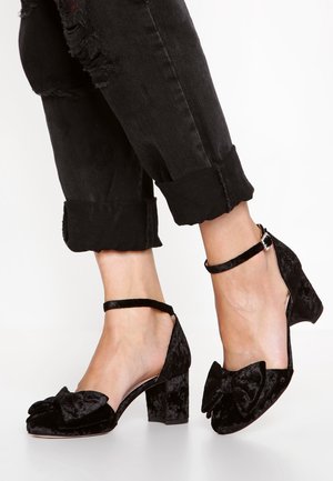 Escarpins à talons blocs en velours noir avec brides à la cheville et grands nœuds sur le devant, portés avec un jean noir déchiré et retroussé.