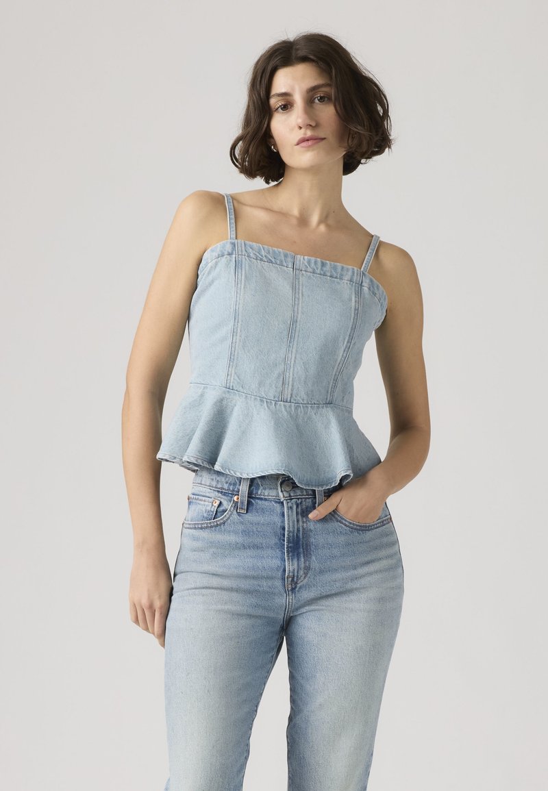 Jonge vrouw met kort bruin haar, gekleed in een lichtblauwe denim peplum top en bijpassende high-waist jeans, staand met één hand in haar zak.