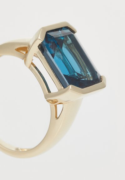 Anillo de oro con una gran piedra azul en corte esmeralda, con acentos geométricos y un diseño de banda abierta. Superficie suave y acabado pulido.
