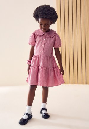 Ung pige med afro hår iført en rødternet kjole, hvide sokker, sorte sko og matchende scrunchie, stående indendørs og kigger ned.
