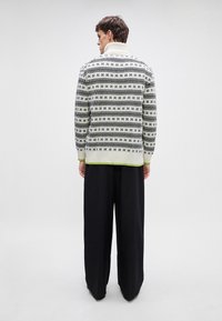 Strikkegenser i svart og hvitt med et geometrisk mønster, med høy krage og grønne detaljer ved mansjettene og kanten; brukt med vide svarte bukser.