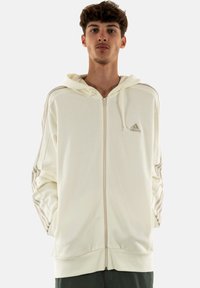 Felpa con zip di colore crema, con polsini e orlo a coste, caratterizzata da tre strisce orizzontali su ciascun manica e un piccolo logo sul petto.