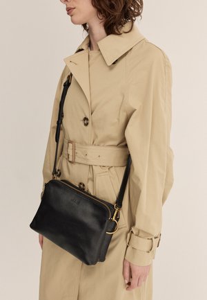 Borsa a tracolla in pelle nera con texture, hardware dorato, chiusura con zip e tracolla regolabile, indossata sopra un trench coat beige.