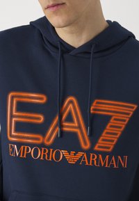 Hoodie di un blu scuro con logo "EA7" arancione luminoso e testo "EMPORIO ARMANI", dotato di tasca a marsupio e cappuccio regolabile con coulisse.