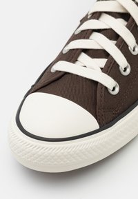 Brun linnesneaker med vit gummistövel, texturerat tyg och gräddfärgade snören. Designad med silveröglor och en svart sulaccent.