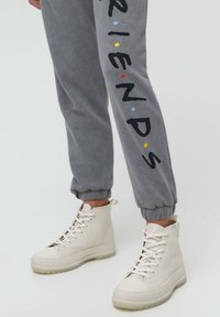 Person iført grå joggingbukser med "PARIS" tekst og hvide høje snøresneakers, der står mod en ensfarvet lys baggrund.